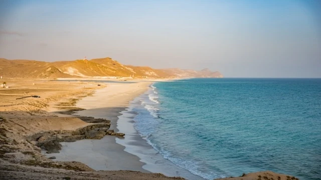 Plage côtière avec du sable blanc s'étendant le long d'une mer bleue et calme, bordée par des collines rocheuses dorées sous une lumière douce de fin de journée. Le paysage offre une vue paisible et dégagée, avec une transition harmonieuse entre terre et mer.