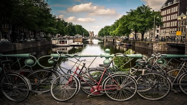 Vue d'un canal emblématique d'Amsterdam avec plusieurs vélos attachés à une rambarde en premier plan, des arbres verdoyants alignés de chaque côté, des bâtiments traditionnels en arrière-plan et un ciel partiellement nuageux au-dessus.