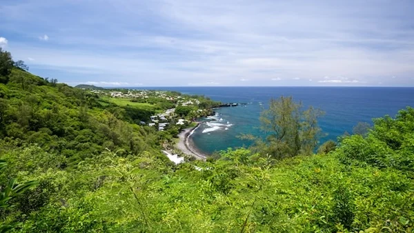 Vue panoramique sur un littoral verdoyant de l'île de la Réunion, avec des collines recouvertes de végétation tropicale, des habitations dispersées le long de la côte et l'océan Indien s'étendant à l'horizon sous un ciel partiellement dégagé.