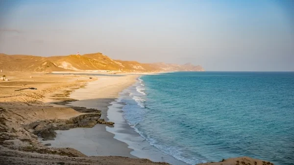 Plage côtière avec du sable blanc s'étendant le long d'une mer bleue et calme, bordée par des collines rocheuses dorées sous une lumière douce de fin de journée. Le paysage offre une vue paisible et dégagée, avec une transition harmonieuse entre terre et mer.